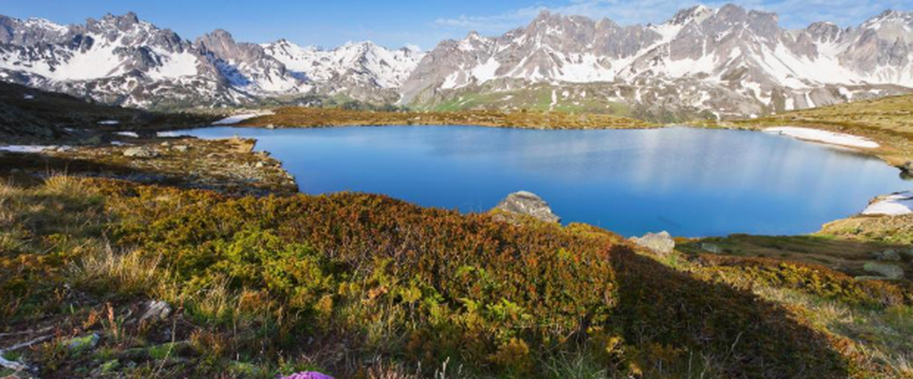 Lac alpin entouré de sommets enneigés et de prairies de montagne dans les Alpes.