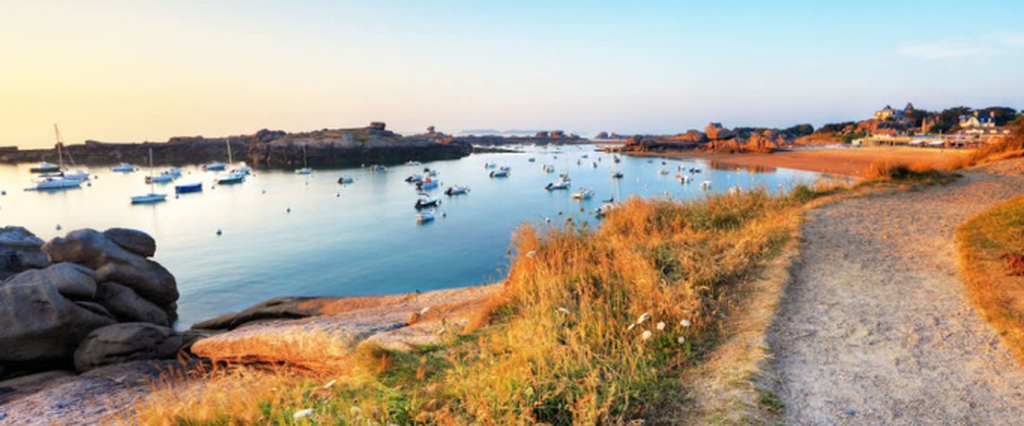 Sentier des Douaniers GR 34 en Bretagne longeant un port côtier avec des bateaux et des rochers.