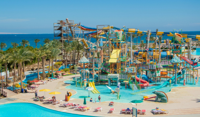 Parc aquatique moderne avec structures de jeux, toboggans multicolores, piscine peu profonde et transats, situé en bord de mer sous un ciel ensoleillé